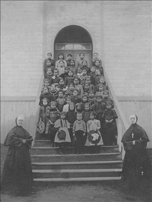 Native American children of different ages stand on the steps of a church. At the bottom of the church are two white nuns. 